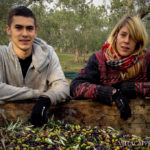 Two people are posing in front of a crate full of olives, showcasing the process of making olive oil.